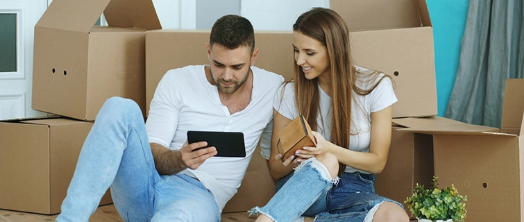 Een glimlachend jong stel zit op de grond tussen kartonnen verhuisdozen; de man houdt een tablet vast terwijl ze elkaar aankijken.